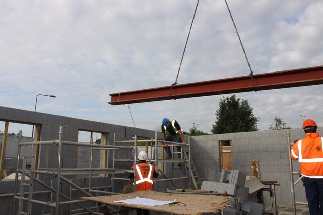 A steel beam suspended from a crane above a completed wall line