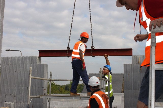 Two workers guiding a steel beam by hand as a crane lowers it into place